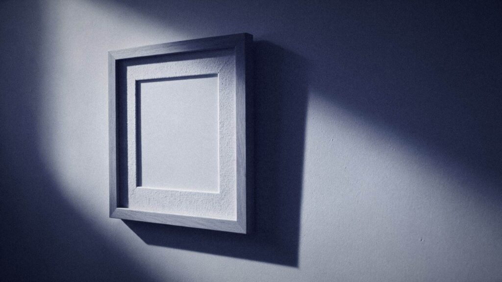 An Empty Picture Frame On A Plain Wall Lit By A Hard Diagonal Shaft Of Light, The Mount Inside Completely Blank